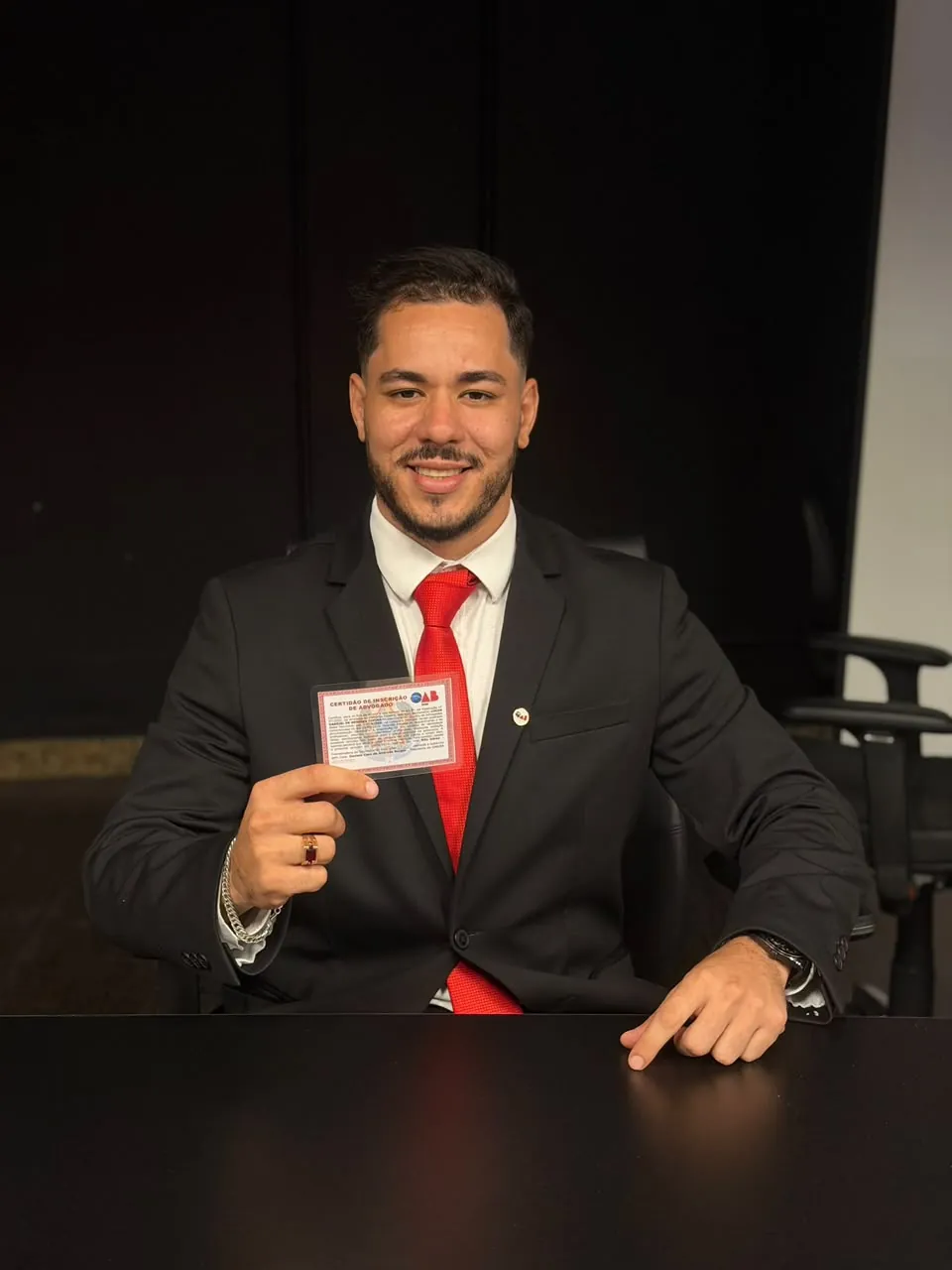 Lucas Tupiná sorrindo, de terno e gravata vermelha, segurando a certidão de inscrição na OAB